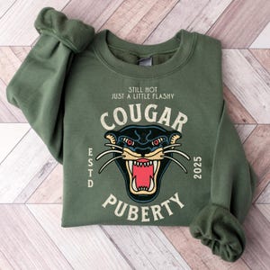 Könnte beinhalten: Olivgrünes Sweatshirt mit dem Text "STILL HOT JUST A LITTLE FLASHY COUGAR PUBERTY 2025" und einer Grafik eines Cougar-Kopfes. Das Sweatshirt hat lange Ärmel und einen Rundhalsausschnitt. Der Text "ESTD" ist ebenfalls aufgedruckt.