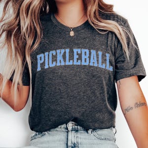 Könnte beinhalten: Dunkelgraues T-Shirt mit dem Wort "PICKLEBALL" in hellblauen Blockbuchstaben auf der Brust. Das T-Shirt mit Rundhalsausschnitt besteht aus weichem Material. Das Design ist lässig und sportlich, geeignet für den Alltag.