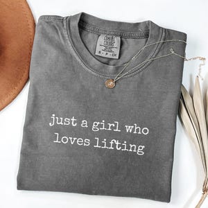 Pode incluir: Uma camiseta cinza Comfort Colors com o texto branco "just a girl who loves lifting". Um colar com pingente dourado e um chapéu marrom também são visíveis.