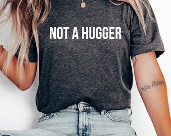 May include: Dark gray t-shirt with the white text "NOT A HUGGER". The shirt is made of a soft material and is a casual wear item. The model is wearing blue jeans.