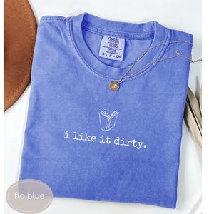 May include: A blue Comfort Colors t-shirt with the text "i like it dirty" and a book graphic. A gold necklace with a pendant rests on the shirt. The shirt is labeled "flo blue".