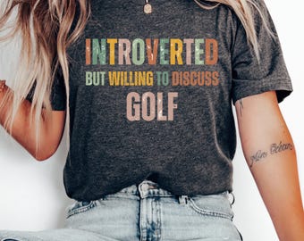Może przedstawiać: Szary t-shirt z napisem "Introvert, ale chętny do dyskusji o golfie" w kolorowych literach. Litery są w retro stylu czcionki.