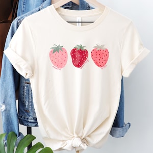 Op de afbeelding: Crèmekleurig T-shirt met drie aardbeienillustraties. Elke aardbei heeft een andere tint roze en rood, met de woorden "freedom", "democracy" en "truth" eronder. Het shirt hangt aan een houten hanger.