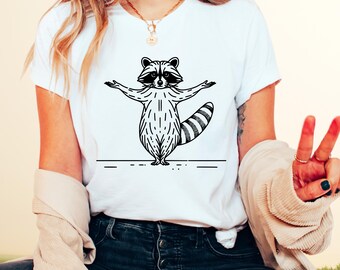 May include: White t-shirt with a black and white illustration of a raccoon balancing on a tightrope. The raccoon has its arms outstretched and its tail is curled up behind it.