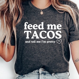以下が含まれることがあります： 「feed me TACOS and tell me I'm pretty」の白文字がプリントされた濃いグレーのTシャツ。クルーネックと半袖です。テキストの下には小さな白いハートがあります。