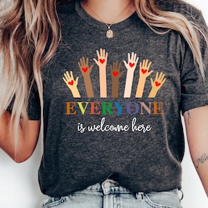 Könnte beinhalten: Dunkelgraues T-Shirt mit dem Text "EVERYONE is welcome here" in Regenbogenfarben. Das Shirt zeigt erhobene Hände verschiedener Hautfarben, jede mit einem roten Herz in der Handfläche.