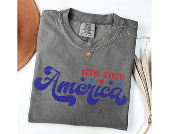 May include: A gray t-shirt with the word "America" in blue script, and the numbers "1776 2026" above it in red. The shirt also has red and blue stars. A gold necklace and a brown hat are also visible.