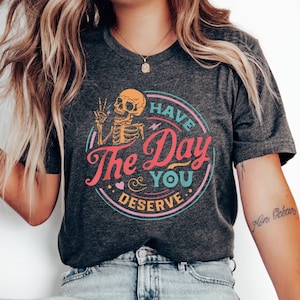 Peut inclure: Un t-shirt gris foncé chiné avec un graphique d'un squelette tenant un signe de paix. Le graphique est entouré d'un cercle avec le texte "Have The Day You Deserve".