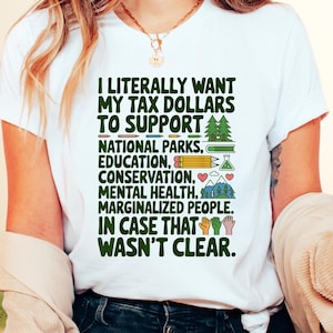 May include: White t-shirt with green text that reads "I literally want my tax dollars to support - National Parks, Education, Conservation, Mental Health, Marginalized People. In case that wasn't clear."