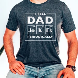 May include: Dark gray t-shirt with the text "I TELL DAD JOKES PERIODICALLY" in a white periodic table-style design. The shirt is made of a soft material and is suitable for casual wear.