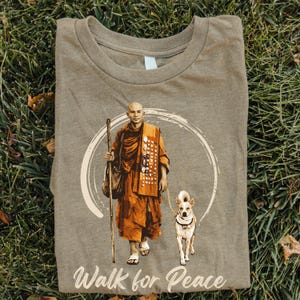 Op de afbeelding: Een heather grijze t-shirt met een afbeelding van een geklede figuur met een hond, lopend in een cirkel. De figuur houdt een staf en een tas vast. De tekst "Walk for Peace" staat onderaan het shirt.