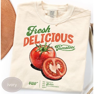 May include: Ivory-colored t-shirt with a graphic of a red tomato and a sliced tomato. The text "Fresh Delicious Tomatoes" is printed above the tomato illustration. The shirt also has the text "No Bigots No Bullies No Billionaires 8647".