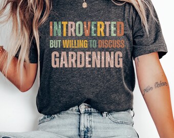 Può includere: Una maglietta grigia scura con il testo "Introverso ma disposto a discutere di giardinaggio" in lettere colorate e consumate.