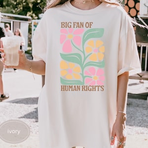 May include: Ivory-colored t-shirt with a floral design in pink, yellow, and green, and the text "BIG FAN OF HUMAN RIGHTS." The shirt is a relaxed fit.