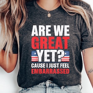 Puede incluir: Una camiseta gris oscuro con el texto "Are We Great Yet? Cause I Just Feel Embarrassed" en blanco con acentos rojos y azules. El texto está en una fuente desgastada y está rodeado por dos pequeños gráficos de banderas americanas.