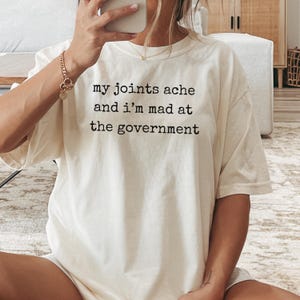 May include: Cream-colored t-shirt with the text "my joints ache and I'm mad at the government" in black. The shirt has a relaxed fit and short sleeves. The image shows the shirt being worn.