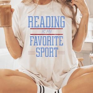 Może przedstawiać: Kremowy t-shirt z napisem "READING IS MY FAVORITE SPORT" w niebieskich literach blokowych. Tekst jest oddzielony czerwonymi liniami. Koszulka jest w stylu casual i wygodnym.