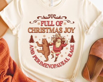 May include: Cream-colored t-shirt with a festive graphic. The design features Santa Claus and a bear dancing, surrounded by snowflakes and Christmas trees. Text reads "FULL OF CHRISTMAS JOY AND PERIMENOPAUSAL RAGE."