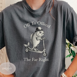 Puede incluir: Camiseta gris oscuro con el texto "Off To Offend The Far Right". El gráfico presenta un pez con bombín montado en un patinete. La camiseta está hecha de un material suave y cómodo.
