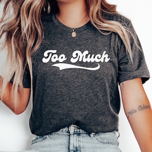 Op de afbeelding: Donkergrijs T-shirt met de witte tekst "Too Much" in een retro lettertype. Het shirt heeft een ronde hals en lijkt gemaakt van een zacht materiaal. De tekst heeft een gebogen onderstreping.