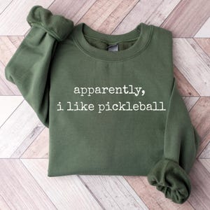 Pode incluir: Um moletom verde-oliva com o texto "apparently, i like pickleball" em branco. O moletom tem mangas compridas e gola redonda. O tecido parece ser um material macio e confortável.