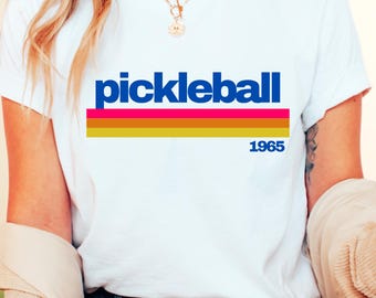 May include: White t-shirt with a blue and pink retro-style graphic that says "pickleball 1965".