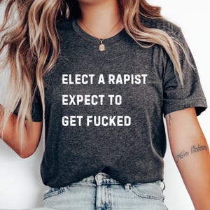 Può includere: T-shirt grigio scuro con la scritta bianca "ELECT A RAPIST EXPECT TO GET FUCKED". La maglietta è a girocollo.