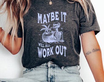 May include: A dark gray t-shirt with a graphic of a skeleton drinking from a coconut. The text on the shirt reads "Maybe it will all work out".