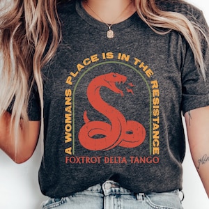 May include: A dark grey t-shirt featuring a graphic of a red snake with the text "A Woman's Place is in the Resistance" in a yellow arch around the snake. The text "Fox Trot Delta Tango" is printed below the snake.