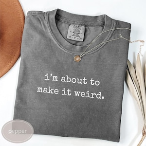 Puede incluir: Una camiseta gris Comfort Colors con la frase "I'm about to make it weird" impresa en blanco. También se ve un collar con colgante dorado y un sombrero marrón. La camiseta está doblada cuidadosamente sobre una superficie blanca.