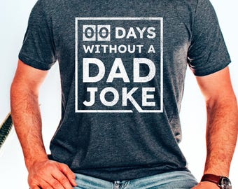 Może przedstawiać: Ciemnoszara koszulka z napisem "00 DAYS WITHOUT A DAD JOKE" w białym prostokącie. Koszulka jest z krótkim rękawem i ma okrągły dekolt. Osoba ma na sobie niebieskie dżinsy i brązowy zegarek.