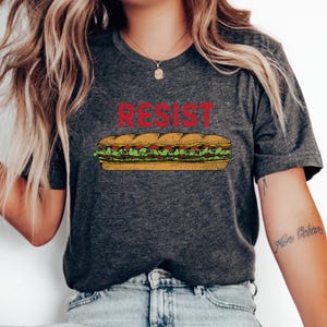 Puede incluir: Camiseta gris oscuro con la palabra "RESIST" en rojo sobre una ilustración de sándwich. El sándwich incluye lechuga, tomates y carne. Una camiseta de estilo casual.