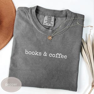 Può includere: Una maglietta grigia con le parole "books & coffee" in corsivo bianco. Una collana con pendente dorato e foglie decorative essiccate sono sulla maglietta. La maglietta è piegata ordinatamente su una superficie bianca. La maglietta è etichettata "pepper".
