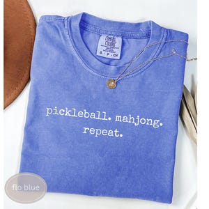 以下が含まれることがあります： ペリウィンクルブルーの半袖クルーネックTシャツ。胸元には「pickleball. mahjong. repeat.」という白い文字がプリントされています。小さな円形のペンダントが付いた細いゴールドトーンのチェーンネックレスが生地の上に置かれています。