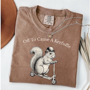 May include: Brown t-shirt with the text "Off To Cause A Kerfuffle" above a squirrel riding a scooter. The squirrel illustration is in black and white. A silver necklace with a small pendant is on the shirt.