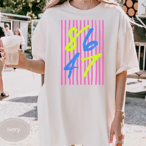 Puede incluir: Camiseta de color marfil con un gráfico rosa neón y verde. El gráfico presenta los números 86 y 47 en una fuente azul y amarilla, sobre un fondo de rayas verticales rosas. La camiseta es de corte holgado.