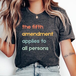 May include: Dark grey t-shirt featuring the text "the fifth amendment applies to all persons" in orange, yellow, teal, and green. The shirt has a crew neck and a casual style.