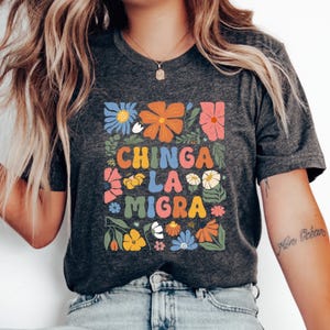 May include: A dark gray t-shirt with a floral design and the text "CHINGA LA MIGRA" in colorful letters.