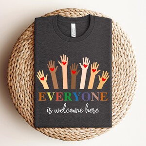Puede incluir: Camiseta gris oscuro con cuello redondo que presenta un diseño de manos levantadas en varios tonos de piel, cada una con un corazón rojo. El texto "EVERYONE is welcome here" está impreso en colores del arcoíris.