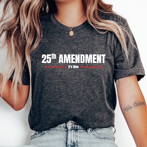 May include: Dark gray t-shirt with the text "25th Amendment" in white, above a line of red stars and the words "it's time". The shirt is a crew neck style.