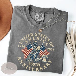 Può includere: Una maglietta grigia Comfort Colors con una grafica che commemora il 250° anniversario degli Stati Uniti d'America. Il design include un'aquila, bandiere americane e la scritta "United States of America 250th Anniversary 1776-2026."