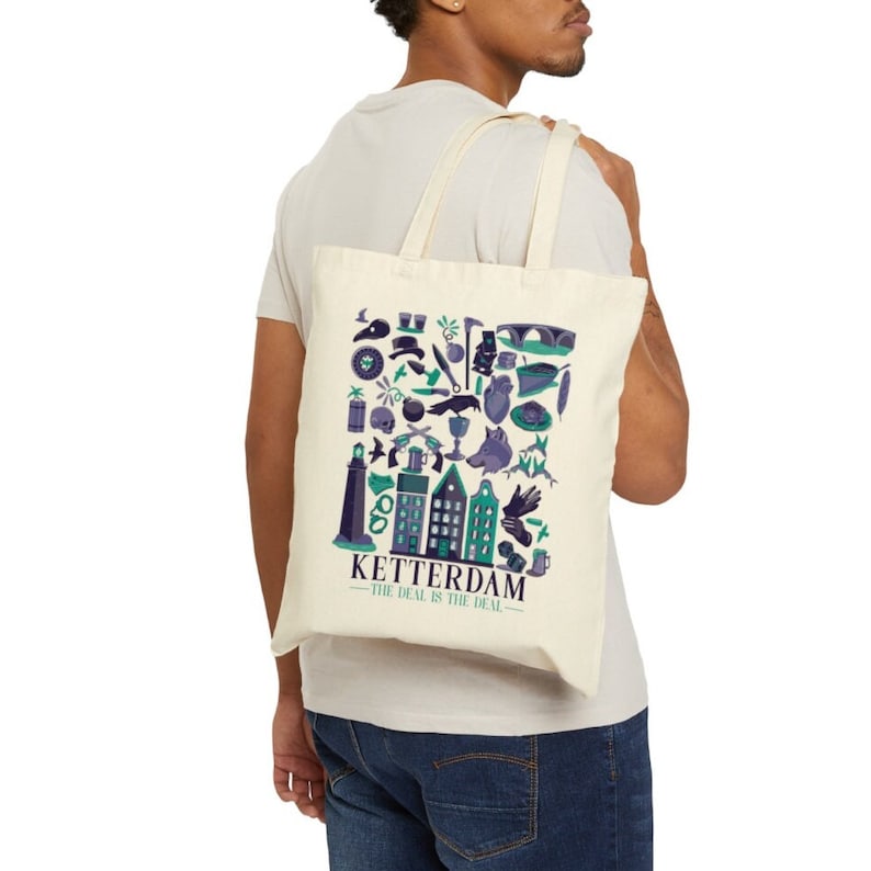 May include: A cream-coloured tote bag with a graphic design in teal, purple, and white. The design features illustrations of buildings, a lighthouse, and other symbols, with the text "KETTERDAM - THE DEAL IS THE DEAL."