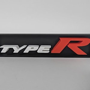 Puede incluir: Letrero rectangular negro con las letras blancas "TYPE" y una "R" roja. Las letras están en relieve y destacan sobre el fondo negro. Probablemente un emblema o decoración de coche.