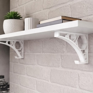 Op de afbeelding: Een witte zwevende plank met decoratieve beugels, gemonteerd op een bakstenen muur. De plank bevat een kleine plant in een pot, een witte keramische container en een stapel boeken. De beugels hebben een geometrisch ontwerp.