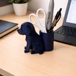 Peut inclure: Un organiseur de bureau bleu marine en forme de chiot assis. Il contient des ciseaux blancs, un marqueur Sharpie gris et un stylo noir. Un ordinateur portable et une petite plante en pot sont en arrière-plan.