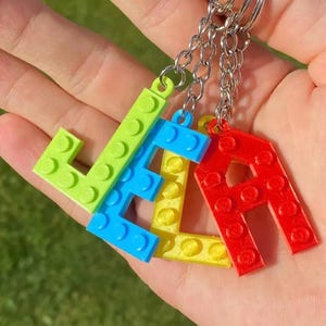 May include: Keychains spelling out the name "JELA" in bright, primary colors. The letters are made to resemble interlocking toy bricks, with a green "J", blue "E", yellow "L", and red "A". Each letter is attached to a silver keyring.