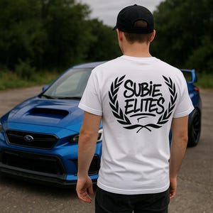 Może przedstawiać: Biała koszulka z napisem "SUBIE ELITES" w czarnym, stylizowanym kroju, obramowana wzorem wieńca laurowego. Niebieski samochód jest widoczny w tle, a także osoba w czarnej czapce.