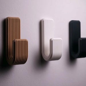 Puede incluir: Tres ganchos de pared con un diseño acanalado, en marrón, blanco y negro. Los ganchos están montados en una pared gris claro. Cada gancho está diseñado para colgar objetos.