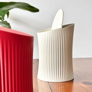 May include: A cream-colored, ribbed vase with a matching lid is partially open, revealing the interior. A red, ribbed vase is in the foreground. Both vases are on a wooden surface, with a green plant in the background.