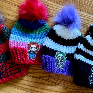 Peut inclure: Quatre bonnets en tricot avec différents personnages de films d'horreur. Les bonnets sont rayés en noir et blanc, rouge et vert, violet et blanc, et noir et blanc. Les personnages sont Freddy Krueger, Pennywise, Chucky et Ghostface.